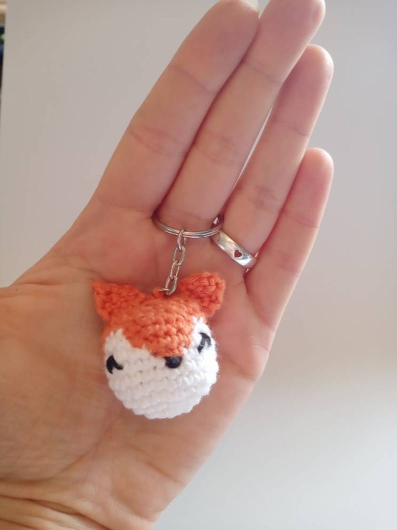 May include: A crocheted fox keychain with orange fur, white face, and black eyes. The keychain is attached to a silver key ring.