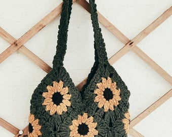 May include: A handmade, dark green crochet bag with a sunflower pattern. The bag features multiple sunflower appliqués in shades of tan and brown. The bag has two long straps for carrying over the shoulder.