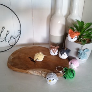 May include: A collection of seven crocheted animal keychains. The keychains are in various colors, including yellow, orange, brown, gray, blue, pink, and green. The keychains are displayed on a wooden surface.