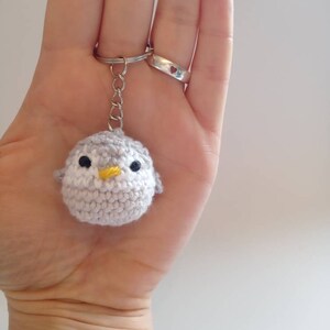 May include: A gray crocheted penguin keychain with a yellow beak and black eyes. The keychain is attached to a silver key ring.