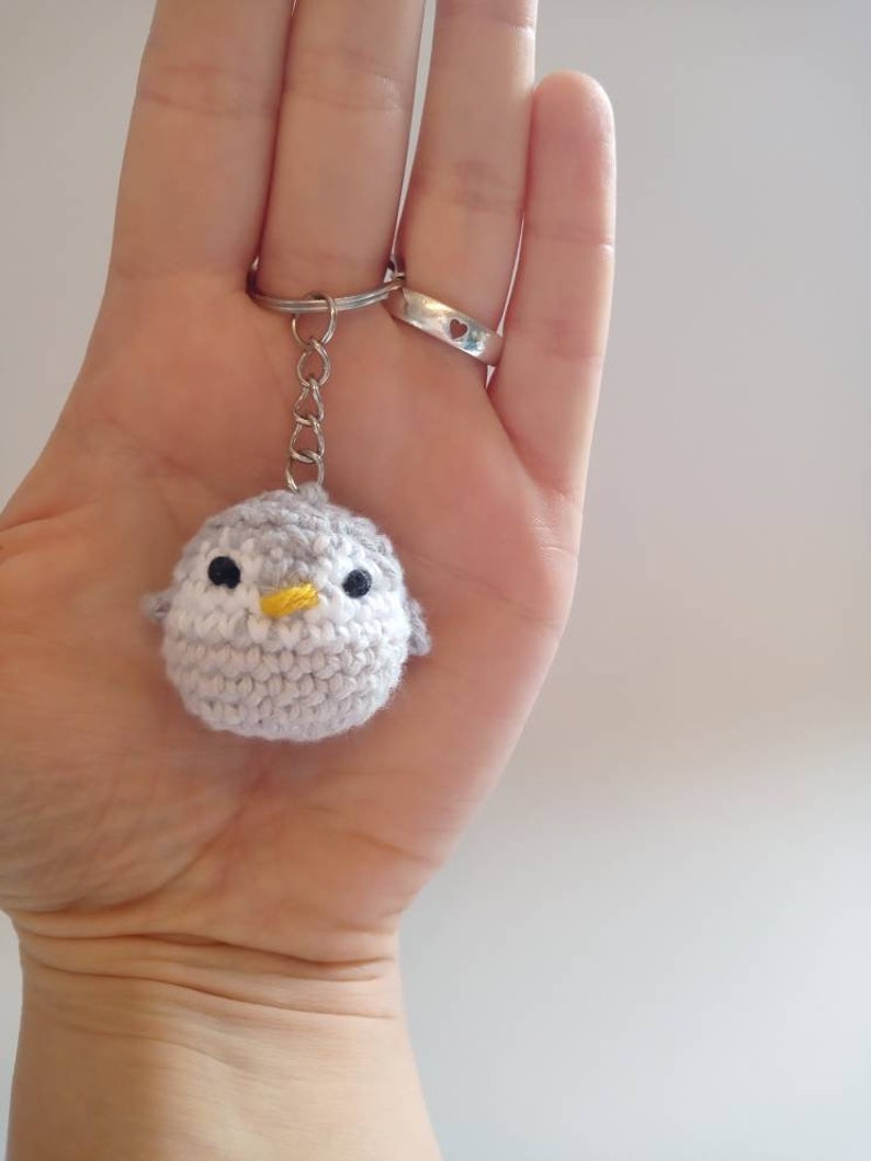 May include: A gray crocheted penguin keychain with a yellow beak and black eyes. The keychain is attached to a silver key ring.