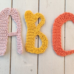 Könnte beinhalten: Gehäkelte Buchstaben A, B und C in rosa, gelb und orange. Die Buchstaben sind horizontal auf einer weißen Holzoberfläche angeordnet.