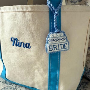 May include: A white canvas tote bag with blue accents, including the handles and a vertical stripe. The name "Nina" is embroidered in blue script. A blue charm with the word "BRIDE" hangs from the handle.