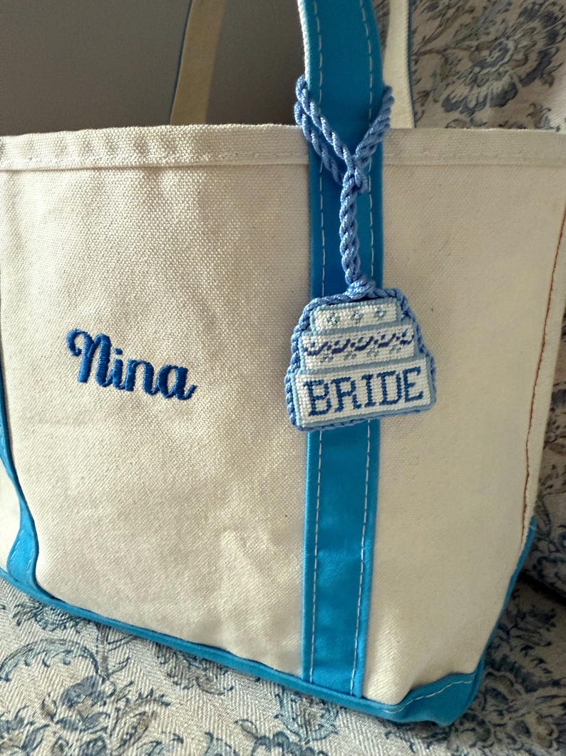 May include: A white canvas tote bag with blue accents, including the handles and a vertical stripe. The name "Nina" is embroidered in blue script. A blue charm with the word "BRIDE" hangs from the handle.