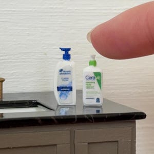 May include: Miniature Head & Shoulders shampoo bottle and CeraVe hydrating facial cleanser bottle on a small bathroom vanity. The shampoo bottle is white with a blue cap, and the cleanser is white and green.