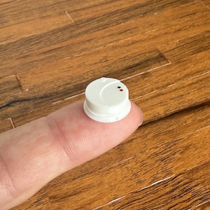 Puede incluir: Un pequeño objeto redondo blanco con un punto rojo y verde en la parte superior, descansando sobre un dedo. El objeto tiene un diseño circular y parece ser un dispositivo en miniatura. El fondo es una superficie de madera.