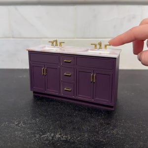 May include: A miniature purple bathroom vanity with two sinks and gold faucets. The countertop is a light pink marble. The vanity has two cabinet doors and three drawers. The background is a white marble tile.