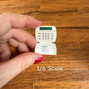 May include: A miniature white security system with a green display and a keypad. The text "1/6 Scale" is printed below the device. The device is held against a wooden background.