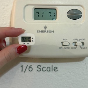 Puede incluir: Un pequeño termostato blanco con una pantalla digital que muestra "72" se sostiene frente a un termostato Emerson más grande. El termostato más grande muestra "71 71". El texto "1/6 Scale" está en la parte inferior de la imagen.
