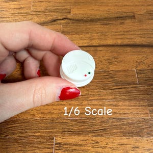 Könnte beinhalten: Ein weißer Miniatur-Rauchmelder mit roter und grüner Kontrollleuchte. Der Text "1/6 Scale" ist darunter aufgedruckt. Dieser Artikel ist wahrscheinlich für ein Puppenhaus oder ein Modell.