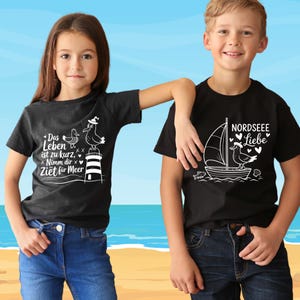 K&ouml;nnte beinhalten: Zwei Kinder in schwarzen T-Shirts mit wei&szlig;en Grafiken. Eines zeigt einen Leuchtturm und einen Vogel mit dem Text "Das Leben ist zu kurz, Nimm dir Zeit f&uuml;r Meer". Das andere zeigt "NORDSEEE Liebe" mit einem Segelboot und Vogel.