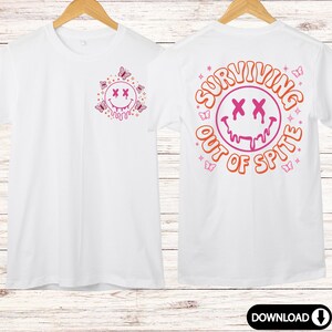 Peut inclure: T-shirt blanc avec un smiley rose et orange aux yeux qui coulent et le texte "SURVIVING OUT OF SPITE" au dos. Le devant du t-shirt pr&eacute;sente un plus petit smiley rose et orange aux yeux qui coulent entour&eacute; de papillons.
