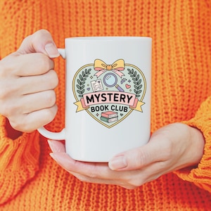 May include: White ceramic mug featuring a heart-shaped design with a magnifying glass, books, and the text "MYSTERY BOOK CLUB." The design includes a bow and laurel leaves. The mug is held by a person wearing an orange sweater.