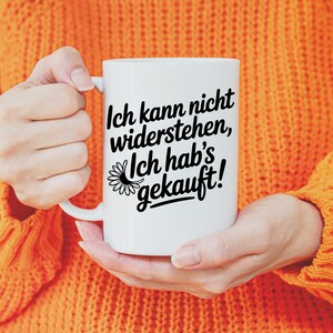 May include: A white ceramic mug with a black inscription that reads "Ich kann nicht widerstehen, Ich hab's gekauft!" and a daisy graphic. The mug is held by a person wearing an orange sweater.
