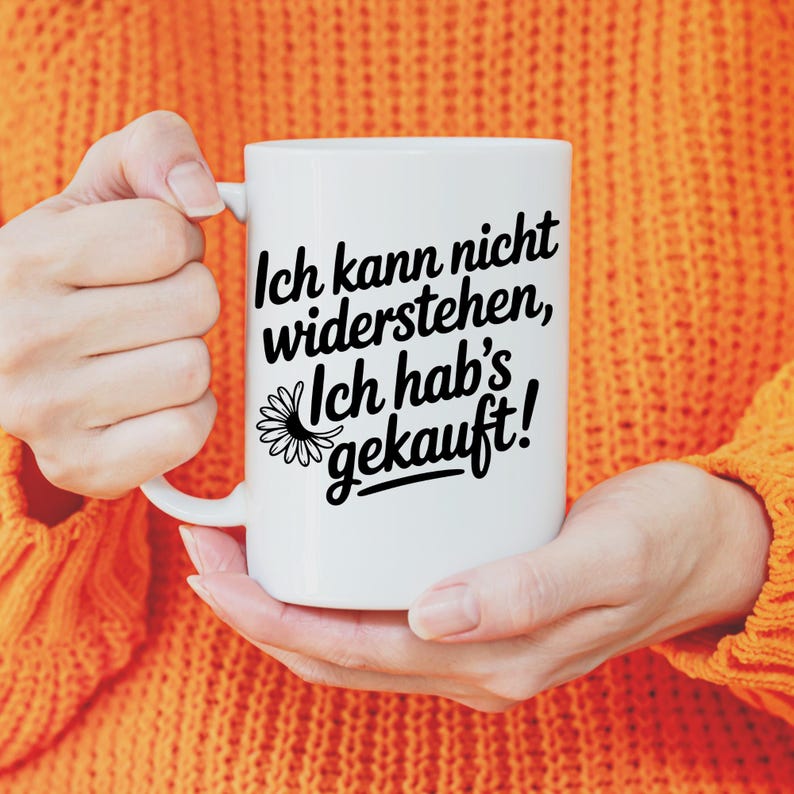 May include: A white ceramic mug with a black inscription that reads "Ich kann nicht widerstehen, Ich hab's gekauft!" and a daisy graphic. The mug is held by a person wearing an orange sweater.