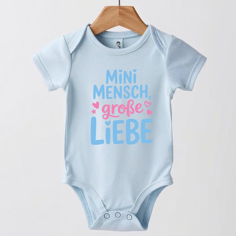 May include: A light blue baby onesie with short sleeves, hanging on a wooden hanger. The onesie has the text "Mini Mensch, gro&szlig;e Liebe" in blue and pink, with a star and heart graphic.