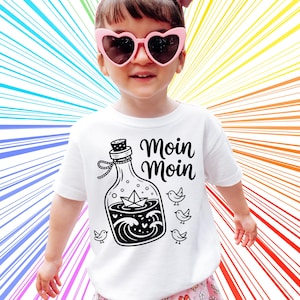 K&ouml;nnte beinhalten: Wei&szlig;es T-Shirt mit einer schwarzen Grafik einer Flasche mit einem Papierboot, Wellen und V&ouml;geln, sowie dem Schriftzug "Moin Moin". Der Hintergrund ist ein farbenfrohes Regenbogenmuster.