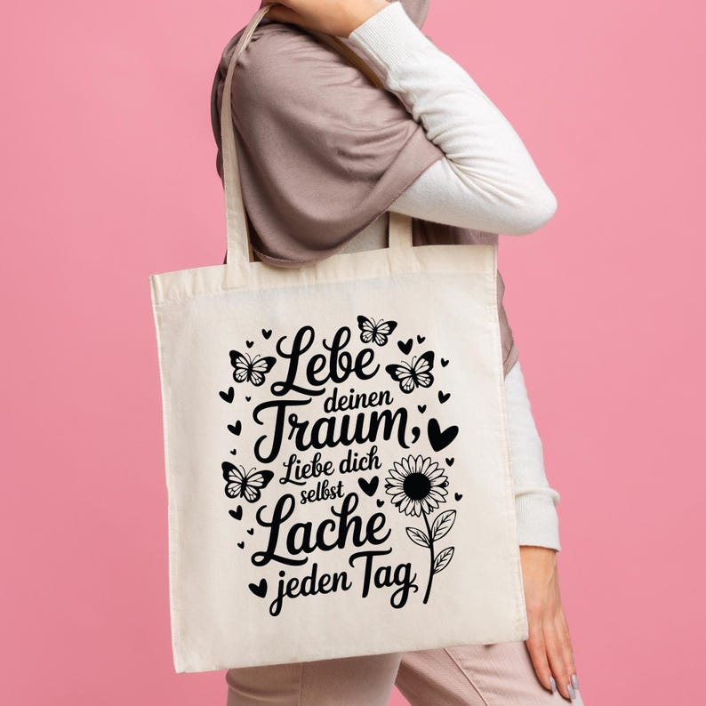 May include: Cream tote bag with a black graphic design featuring the words "Lebe deinen Traum" and "Lache jeden Tag". The design includes butterflies, hearts, and a sunflower. The bag is held by a person.