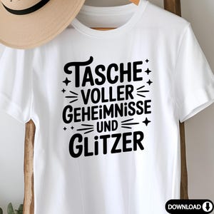 Puede incluir: Camiseta blanca con el texto alem&aacute;n "TASCHE VOLLER GEHEIMNISSE UND GLITZER" en negro. El dise&ntilde;o incluye estrellas y l&iacute;neas. La camiseta se muestra en una percha de madera.
