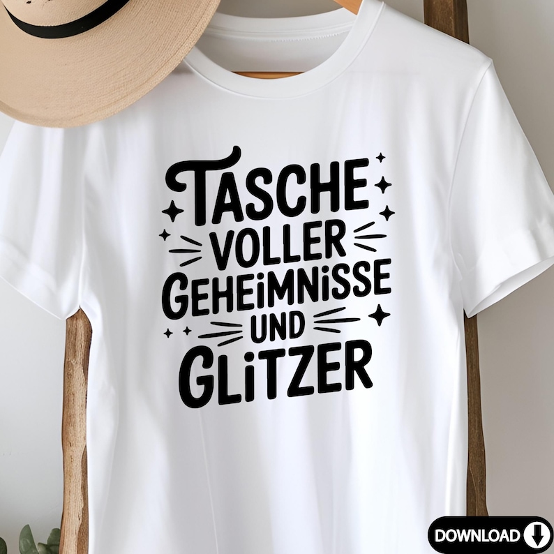 Puede incluir: Camiseta blanca con el texto alem&aacute;n "TASCHE VOLLER GEHEIMNISSE UND GLITZER" en negro. El dise&ntilde;o incluye estrellas y l&iacute;neas. La camiseta se muestra en una percha de madera.