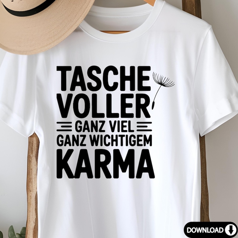 May include: White t-shirt with the German text "TASCHE VOLLER = GANZ VIEL = GANZ WICHTIGEM KARMA" in black, along with a dandelion graphic. The shirt is hanging on a wooden rack.