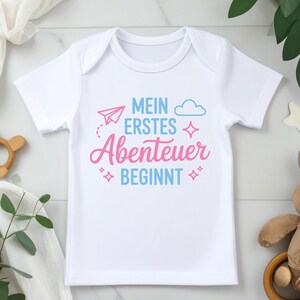 May include: White baby onesie with the German text "MEIN ERSTES ABENTEUER BEGINNT" in blue and pink. The design includes a paper airplane, cloud, and star accents. A cute and stylish outfit for a newborn.