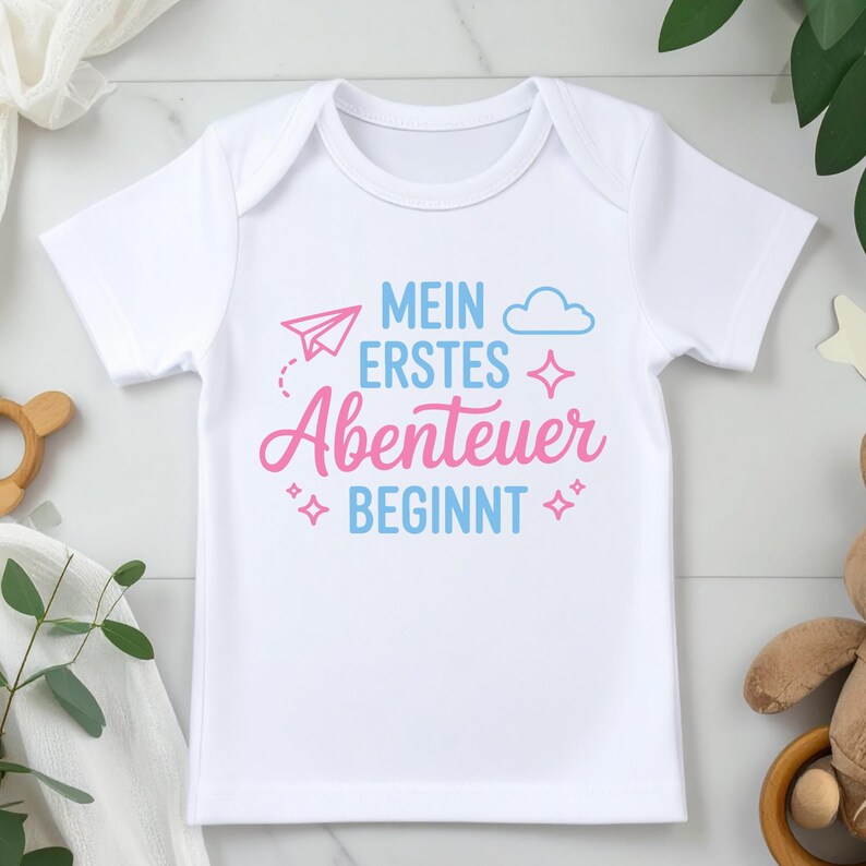 May include: White baby onesie with the German text "MEIN ERSTES ABENTEUER BEGINNT" in blue and pink. The design includes a paper airplane, cloud, and star accents. A cute and stylish outfit for a newborn.