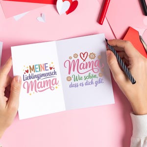 May include: A white greeting card with the word "Mama" in pink script, decorated with hearts and flowers. The card is open, revealing text in German. A hand holds a black and silver pen, ready to write.