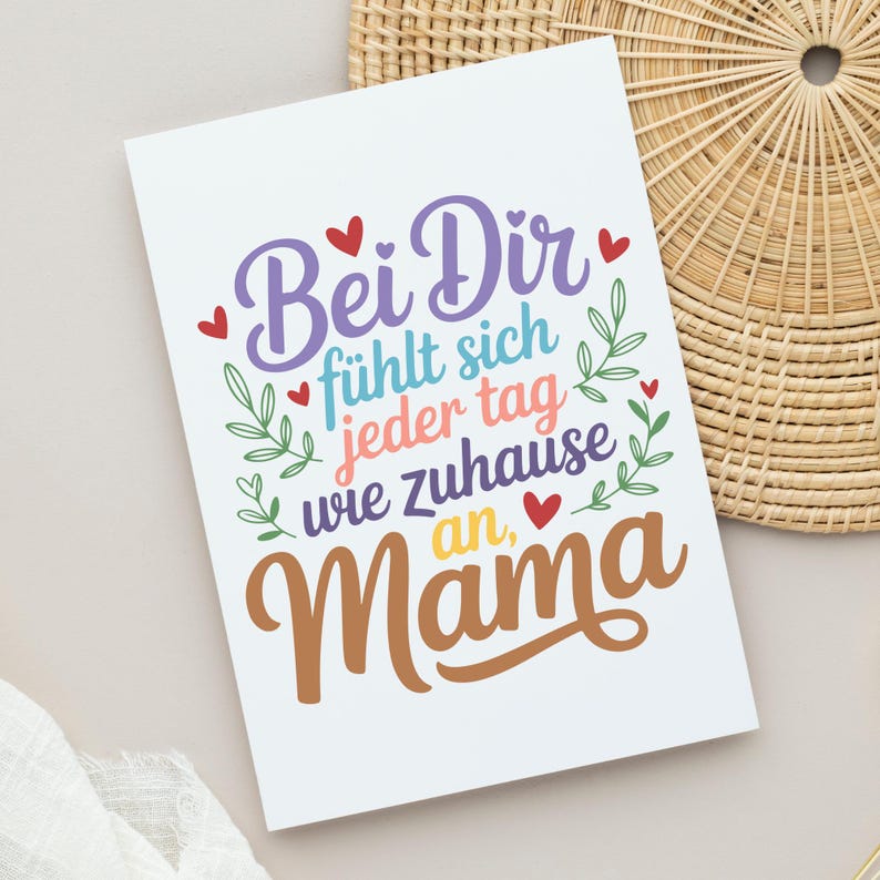 May include: A white greeting card with the German text "Bei Dir f&uuml;hlt sich jeder Tag wie zuhause an, Mama" in colorful script. The design includes red hearts and green leaf accents, perfect for Mother's Day or a special occasion.