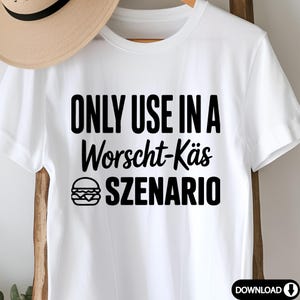 May include: White t-shirt with the text "ONLY USE IN A Worscht-K&auml;s SZENARIO" in black, along with a burger graphic. The shirt is hanging on a wooden rack. A straw hat is partially visible in the background.