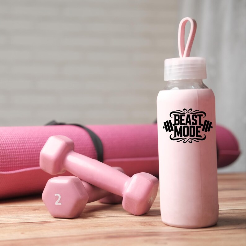Może przedstawiać: R&oacute;żowa butelka na wodę z czarnym nadrukiem "Beast Mode" i r&oacute;żową silikonową osłoną. Dwa r&oacute;żowe hantle na pierwszym planie.