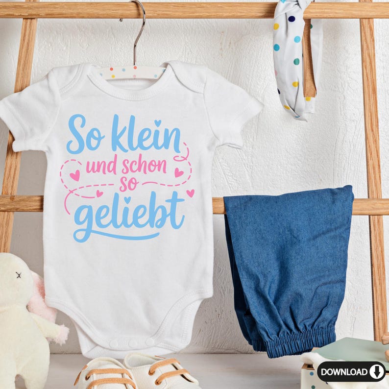 May include: A white baby onesie with the text "So klein und schon so geliebt" in blue and pink. The onesie is hanging on a wooden rack with a pair of blue pants, a headband, and baby shoes. A stuffed bunny is also visible.