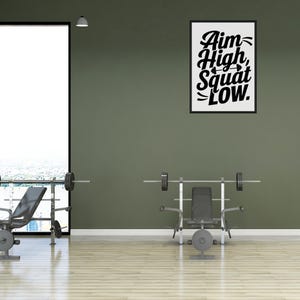 Może przedstawiać: Motywujące zdanie "Aim High, Squat Low" jest wyświetlane w czarnej ramie na zielonej ścianie. Na pierwszym planie znajdują się dwie ławki do ćwiczeń z sztangami.