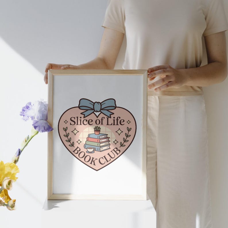 May include: A framed print featuring a heart-shaped design with the text "Slice of Life Book Club." The design includes a stack of books, a teacup, and a bow. The print is held by a person.