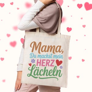 May include: A beige tote bag with the words "Mama, Du machst mein Herz L&auml;cheln" in brown, blue, pink, and green. The bag has a floral and heart design. The bag is being held by a person.