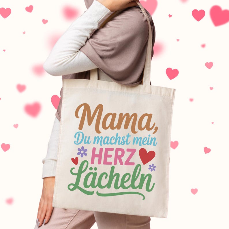 May include: A beige tote bag with the words "Mama, Du machst mein Herz L&auml;cheln" in brown, blue, pink, and green. The bag has a floral and heart design. The bag is being held by a person.