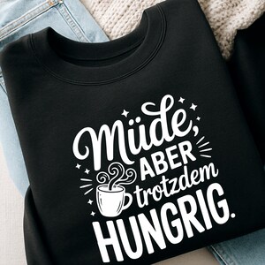 Puede incluir: Sudadera negra con texto blanco "M&uuml;de, aber trotzdem hungrig". Se incluye un gr&aacute;fico de taza de caf&eacute; en el dise&ntilde;o. La sudadera se muestra sobre una superficie de color claro.