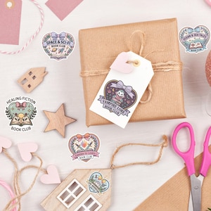 May include: A wrapped brown gift box with a white tag featuring a heart-shaped design and the text "VAMPIRE LORE BOOK CLUB." Scattered around are stickers with book club themes, including "SPACE & SCI-FI," "HEALING FICTION," and "Enemies to Lovers."