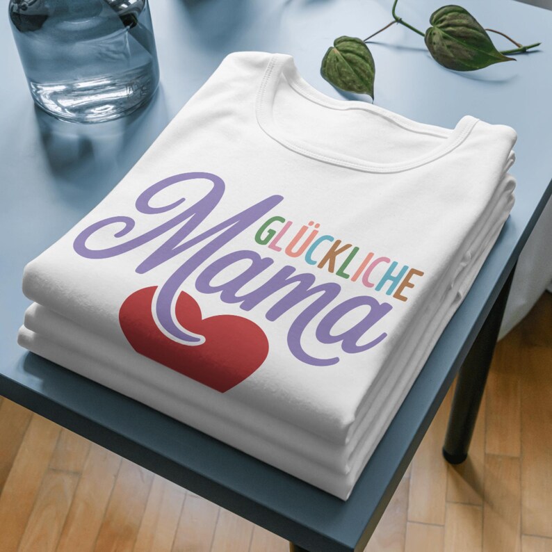May include: A stack of white t-shirts with the word "Mama" in purple script, a red heart, and the words "GL&Uuml;CKLICHE" in colorful letters. The shirts are neatly folded on a blue surface.