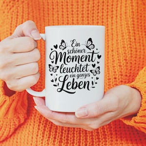 May include: White ceramic mug with a black design featuring butterflies, hearts, and the German text "Ein sch&ouml;ner Moment leuchtet ein ganzes Leben." The mug is held by a person wearing an orange jumper.