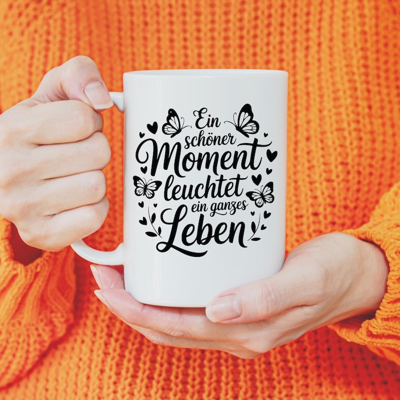 May include: White ceramic mug with a black design featuring butterflies, hearts, and the German text "Ein sch&ouml;ner Moment leuchtet ein ganzes Leben." The mug is held by a person wearing an orange jumper.