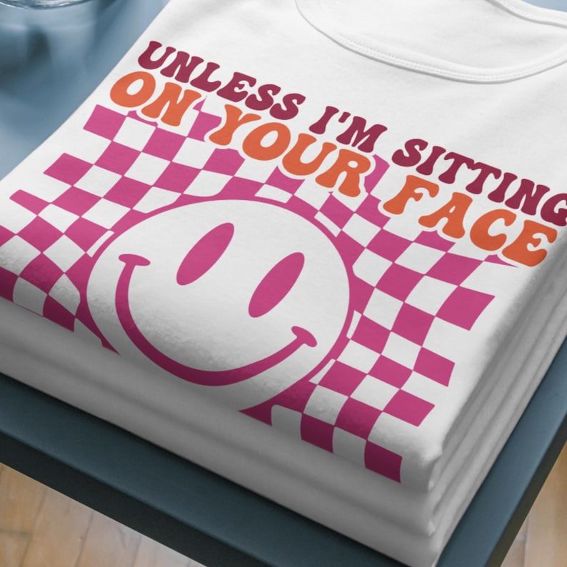 Peut inclure: T-shirt blanc avec un motif &agrave; carreaux rose et blanc et un smiley rose. Le texte "Unless I'm sitting on your face" est &eacute;crit en lettres orange et rose.