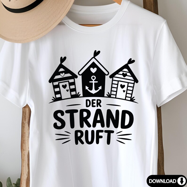 K&ouml;nnte beinhalten: Wei&szlig;es T-Shirt mit einem schwarzen Aufdruck von drei Strandh&uuml;tten, Herzen und einem Anker. Der deutsche Text "DER STRAND RUFT" ist darunter gedruckt. Ein Strohhut ist teilweise im Hintergrund zu sehen.