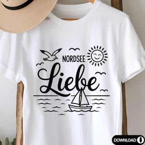 K&ouml;nnte beinhalten: Wei&szlig;es T-Shirt mit schwarzer Grafik. Das Design zeigt das Wort "Liebe" &uuml;ber einem Segelboot auf dem Wasser, eine Sonne, eine M&ouml;we und die Worte "Nordsee". Ein Strohhut ist teilweise sichtbar.