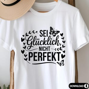 May include: White t-shirt featuring the German text "Sei Gl&uuml;cklich, Nicht Perfekt", translating to "Be Happy, Not Perfect". The design includes black butterflies and heart-shaped accents.