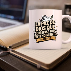 May include: White ceramic mug with a handle, featuring the text "LA PAZ DE DIOS QUE SOBREPASA TODO ENTENDIMIENTO" and "-FILIPENSES 4:7-". The design includes a white dove with an olive branch. The mug is on a notebook with a pen.
