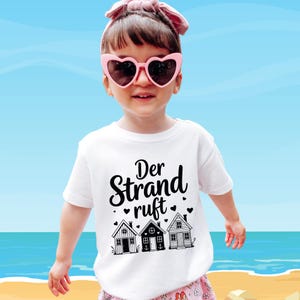 K&ouml;nnte beinhalten: Ein wei&szlig;es T-Shirt mit dem deutschen Text "Der Strand ruft" und drei kleinen Hausillustrationen. Das Kind tr&auml;gt eine rosa herzf&ouml;rmige Sonnenbrille und ein rosa Stirnband. Der Hintergrund zeigt einen blauen Himmel und das Meer.
