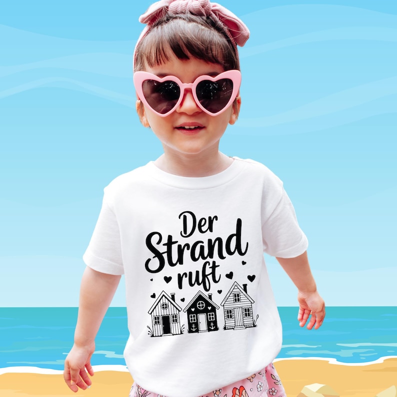 K&ouml;nnte beinhalten: Ein wei&szlig;es T-Shirt mit dem deutschen Text "Der Strand ruft" und drei kleinen Hausillustrationen. Das Kind tr&auml;gt eine rosa herzf&ouml;rmige Sonnenbrille und ein rosa Stirnband. Der Hintergrund zeigt einen blauen Himmel und das Meer.