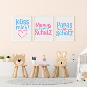 May include: Three framed prints with German text in a child's room. The prints say "K&uuml;ss mich!" with a pink heart, "Mamas Schatz" and "Papas Schatz" with arrows and hearts. The room has bear and bunny chairs.