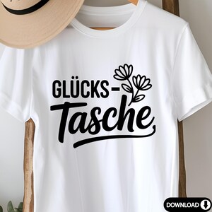 May include: White t-shirt featuring the German text "GL&Uuml;CKS-Tasche" in black, with a floral illustration. The shirt is made of a soft material and is suitable for casual wear. A straw hat is partially visible in the background.